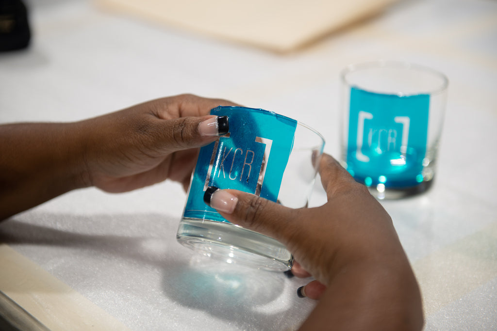 A woman's hands pictured, applicating a blue photo resistant mask onto a whiskey glass prior to engraving. 